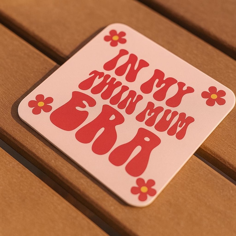 Puede incluir: Un posavasos cuadrado de color rosa claro con la frase "IN MY TWIN MUM ERA" en rojo, con un estilo retro. Peque&ntilde;os gr&aacute;ficos de flores rojas se colocan alrededor de los bordes. El posavasos est&aacute; sobre una superficie de madera.