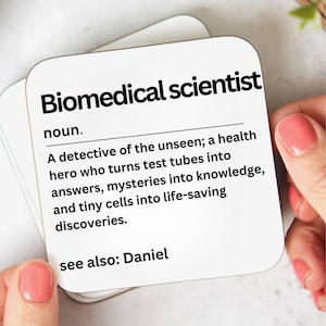 May include: White square coaster with the text "Biomedical scientist" in black. The definition of a biomedical scientist is printed below the title. The text is held by a person with pink nail polish.