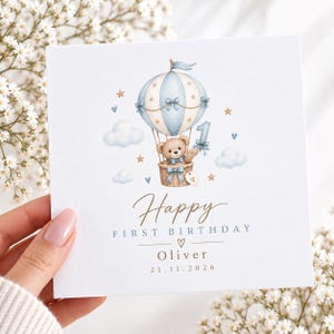 Puede incluir: Una tarjeta de felicitación cuadrada con una ilustración en acuarela de un osito de peluche en un globo aerostático azul y blanco con el número uno, nubes y estrellas. El texto dice Happy FIRST BIRTHDAY, Oliver, 21.11.2026.