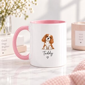 May include: A white ceramic mug with a pink handle and interior rim. The front features a brown and white Cavalier King Charles Spaniel illustration above the name Teddy written in black script, followed by a small black heart symbol.