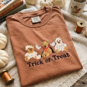 May include: A rust-colored crewneck t-shirt featuring an embroidered design of three teddy bears in Halloween costumes, including a mummy, a vampire, and a ghost, with the black embroidered text Trick or Treat below the bears.