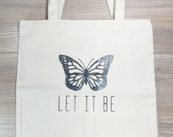 May include: A white canvas tote bag with black handles and a black butterfly design. The text "LET IT BE" is printed below the butterfly.