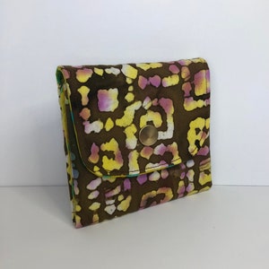 May include: A small brown fabric wallet with a yellow, pink, and white geometric pattern. The wallet has a flap closure with a gold button.