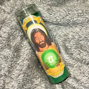 Könnte beinhalten: Eine grüne Glas-Kerze mit dem Bild einer Person mit langen braunen Haaren und Bart, die eine grüne Kugel mit einem Symbol hält. Die Kerze hat ein gold-grünes Etikett, vor einem grau strukturierten Hintergrund.