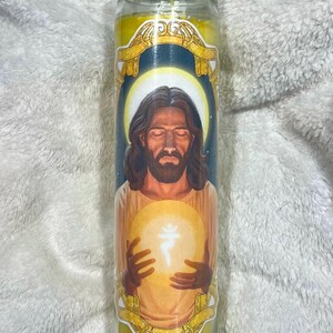 Könnte beinhalten: Eine gelbe Glaskerze mit einem Bild von Jesus, der eine leuchtende Kugel hält. Die Kerze hat einen goldenen Rand und einen dunkelblauen Hintergrund.
