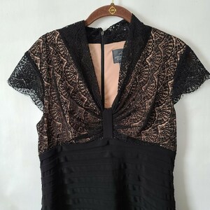 Puede incluir: Vestido negro con corpi&ntilde;o de encaje y mangas cortas. El corpi&ntilde;o presenta intrincados detalles de encaje sobre un forro nude. El vestido tiene un cuello alto y una falda plisada. La marca "Adrianna Papell" es visible en una etiqueta.