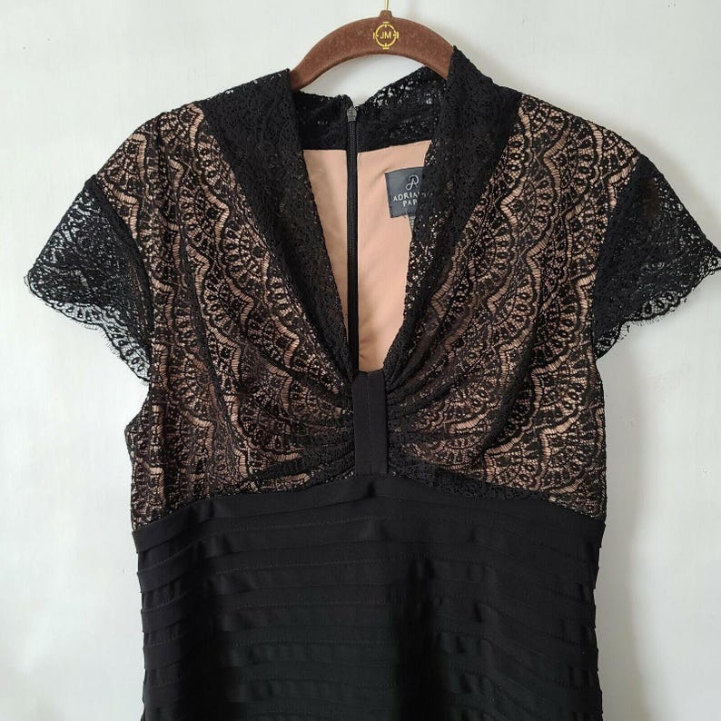 Puede incluir: Vestido negro con corpi&ntilde;o de encaje y mangas cortas. El corpi&ntilde;o presenta intrincados detalles de encaje sobre un forro nude. El vestido tiene un cuello alto y una falda plisada. La marca "Adrianna Papell" es visible en una etiqueta.
