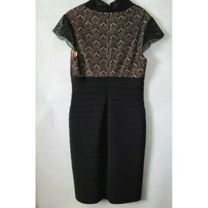 Puede incluir: Vestido negro ajustado con cuello alto y mangas cortas. El corpi&ntilde;o presenta una superposici&oacute;n de encaje beige con un patr&oacute;n festoneado, mientras que la falda tiene una textura acanalada. El vestido es de largo hasta la rodilla.