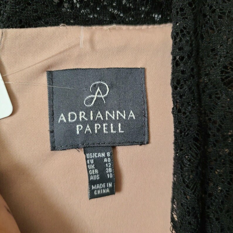 Puede incluir: Primer plano de una etiqueta negra con el nombre de la marca "ADRIANNA PAPELL" en letras blancas. La etiqueta incluye conversiones de tallas para tallas de EE. UU., UE, Reino Unido, GER y AUS. La etiqueta est&aacute; cosida en una prenda de encaje negro.