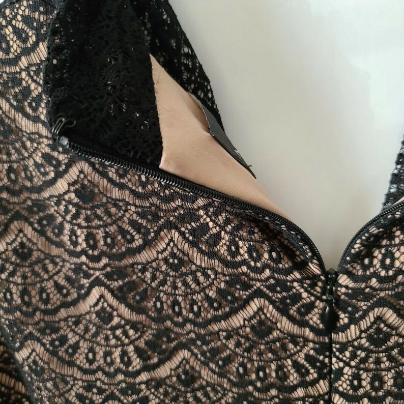 Puede incluir: Primer plano de una prenda de encaje negro con forro beige. El intrincado patr&oacute;n de encaje es visible, y una cremallera recorre la parte delantera. La prenda parece ser una blusa o un vestido, mostrando un dise&ntilde;o delicado y elegante.