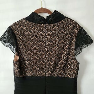 Puede incluir: Vestido negro con cuello alto y mangas cortas. El corpi&ntilde;o presenta una capa inferior beige con una superposici&oacute;n de encaje negro. Las mangas y el escote est&aacute;n ribeteados con encaje negro. El vestido est&aacute; colgado de una percha de madera.