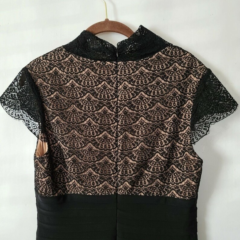 Puede incluir: Vestido negro con cuello alto y mangas cortas. El corpi&ntilde;o presenta una capa inferior beige con una superposici&oacute;n de encaje negro. Las mangas y el escote est&aacute;n ribeteados con encaje negro. El vestido est&aacute; colgado de una percha de madera.