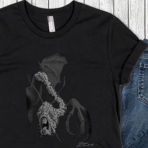 May include: Black t-shirt featuring a detailed grayscale illustration of a creature with a wing and claw. The design is composed of intricate patterns and text. The shirt is paired with blue jeans.