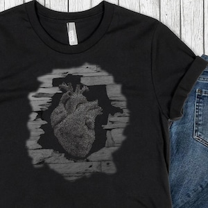May include: Black t-shirt featuring a detailed, textured heart design within a distressed, rectangular frame. The heart appears to be made of small, individual elements. The shirt is paired with blue denim jeans.
