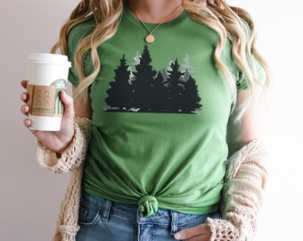 May include: A green t-shirt with a black silhouette of a forest on the front. The shirt is tied at the waist and the person wearing it is holding a coffee cup.