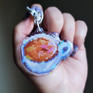 May include: A clear iridescent keychain featuring a pixelated image of a brown bowl of food. The keychain is held in a hand.