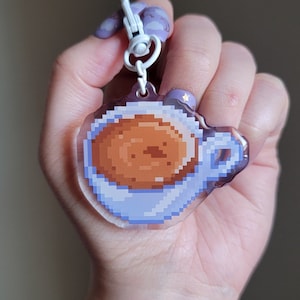May include: A clear acrylic keychain featuring a pixelated design of a blue coffee cup filled with brown coffee. The keychain is attached to a white and silver chain.