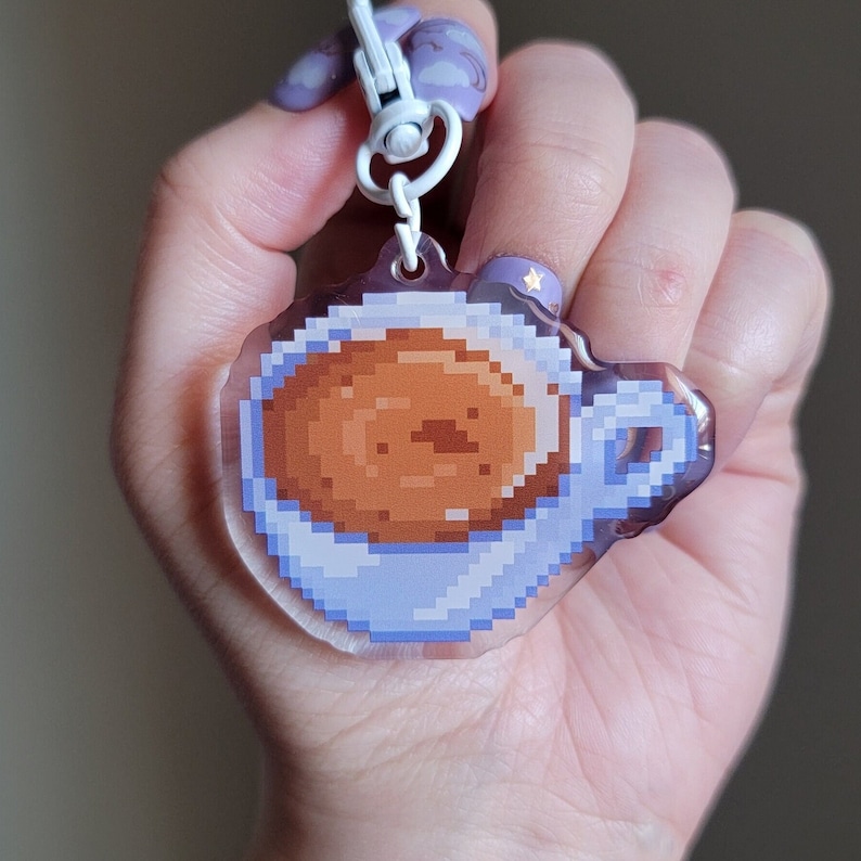 May include: A clear acrylic keychain featuring a pixelated design of a blue coffee cup filled with brown coffee. The keychain is attached to a white and silver chain.