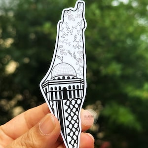 May include: A black and white sticker of the Dome of the Rock in Jerusalem, with a floral pattern behind it. The sticker is shaped like the outline of Israel. The sticker is held in a hand.