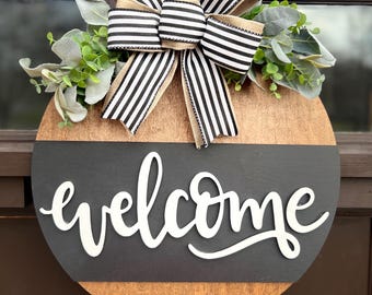 May include: A round wooden door hanger with a black and white striped bow and greenery. The bottom half of the door hanger is black with the word "welcome" written in white.