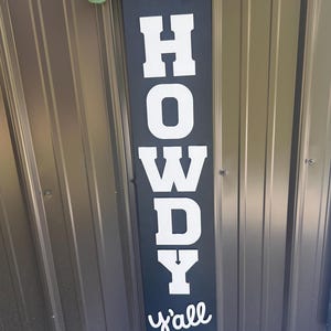 May include: A black wooden sign with the word "HOWDY y'all" in white lettering, and a Highland cow illustration. The sign is decorated with a buffalo plaid bow, burlap ribbon, and faux greenery.
