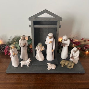May include: A Nativity scene with figures of Mary, Joseph, baby Jesus, shepherds, and animals. The figures are in white robes and are arranged in front of a wooden stable. The base is a dark gray color.