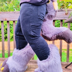 May include: A person wearing black jeans and furry, gray and purple costume feet with yellow claws. A matching tail is visible. The costume is on a wooden deck with a green background.