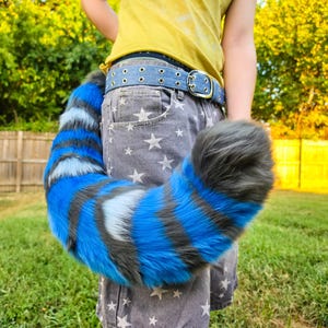 May include: A long, plush, faux fur tail featuring alternating horizontal stripes of vibrant blue, light blue, and dark charcoal gray. The tail is attached to a blue denim belt with silver-toned metal grommets and a rectangular buckle.