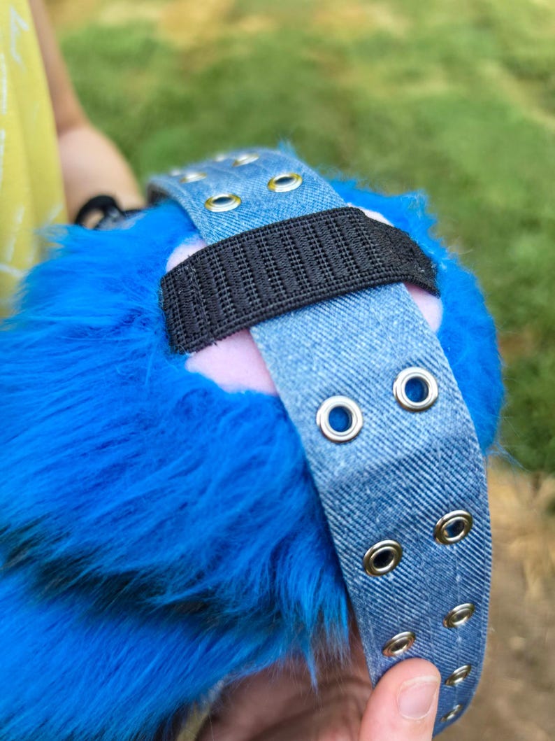 May include: A bright blue faux fur accessory featuring a light blue denim strap with silver-toned metal grommets. A black elastic band is attached to the denim strap, which rests against a soft pink base visible beneath the blue fur texture.