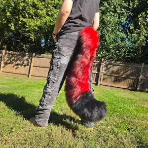 May include: A long, plush faux fur tail featuring a vibrant gradient pattern that transitions from a bright red base at the top to a deep black tip. The tail is attached to the back of grey denim cargo pants worn with a plain black t-shirt.