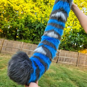 May include: A long, plush faux fur tail featuring a striped pattern of vibrant blue, dark gray, and white bands. The tail has a thick, rounded end and tapers slightly toward the top, showcasing a soft, textured appearance with multi-colored synthetic fibers.