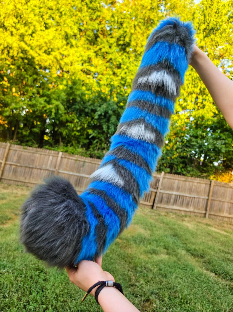 May include: A long, plush faux fur tail featuring a striped pattern of vibrant blue, dark gray, and white bands. The tail has a thick, rounded end and tapers slightly toward the top, showcasing a soft, textured appearance with multi-colored synthetic fibers.