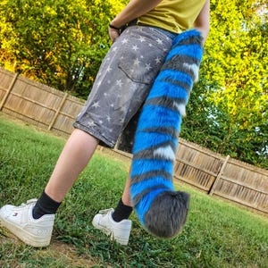 May include: A long, plush faux fur tail featuring alternating horizontal stripes of vibrant blue, dark gray, and white. The tail has a thick, fluffy texture and a rounded dark gray tip, designed to be worn attached to the back of gray star-print shorts.