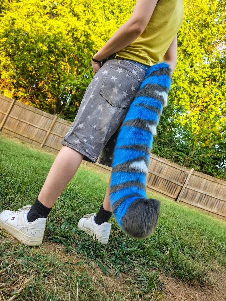 May include: A long, plush faux fur tail featuring alternating horizontal stripes of vibrant blue, dark gray, and white. The tail has a thick, fluffy texture and a rounded dark gray tip, designed to be worn attached to the back of gray star-print shorts.