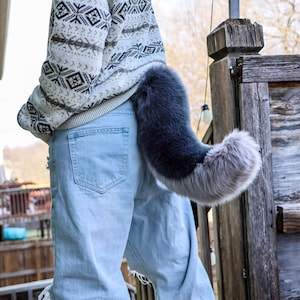 May include: A gray and silver faux fur tail attached to the back of light blue jeans. The tail is curved and fluffy, with a darker gray color at the base that fades to silver at the tip. The person is wearing a patterned sweater.