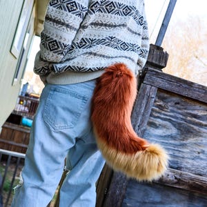 May include: A faux fur tail in shades of orange and tan, attached to the back of a person wearing light blue jeans. The tail is fluffy and curved, with a darker orange top and a lighter tan tip. The person is wearing a cream-colored sweater.