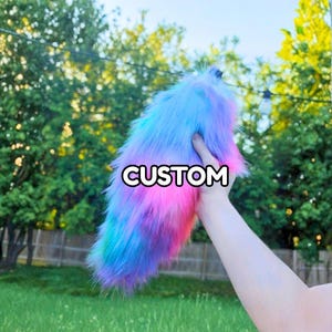 May include: A fluffy, long-haired faux fur tail accessory featuring a vibrant gradient of blue, purple, and pink colors. The word CUSTOM is printed in white capital letters across the center of the plush, textured surface of the tail.