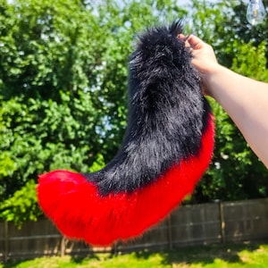 May include: A long, curved faux fur tail accessory featuring a thick layer of black fur on the upper portion that transitions into a vibrant, solid red fur tip. The plush, textured material is shaped into a tapered, animal-like tail design.