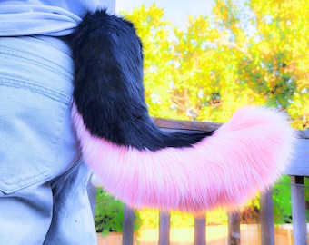 May include: A black and pink faux fur tail, attached to the back of a person's pants. The tail is black at the top and transitions to a vibrant pink at the tip. The tail is fluffy and curved, with a soft texture.