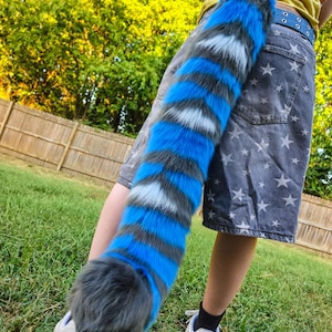 May include: A long, plush faux fur tail featuring alternating horizontal stripes in vibrant blue, dark gray, and white. The tail has a thick, rounded tip and is attached to a pair of gray denim shorts patterned with white stars and a blue belt.