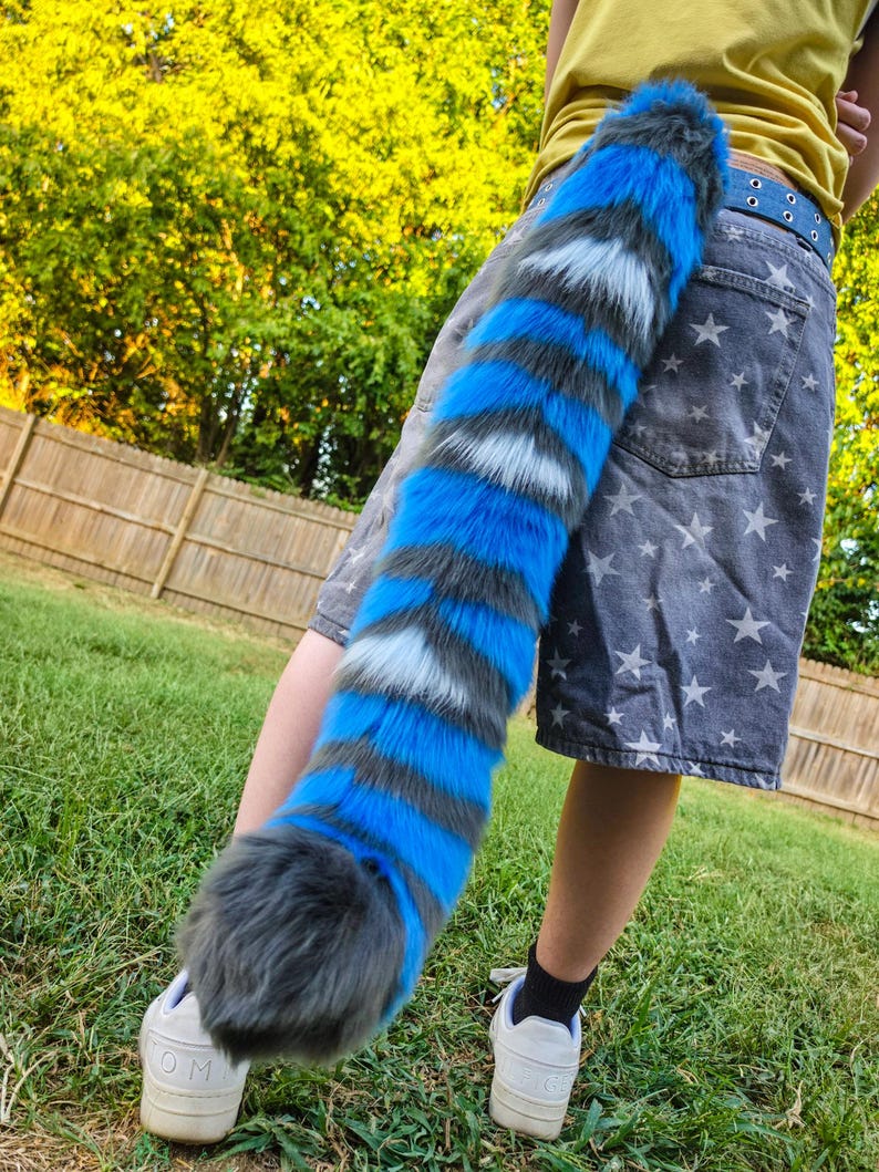 May include: A long, plush faux fur tail featuring alternating horizontal stripes in vibrant blue, dark gray, and white. The tail has a thick, rounded tip and is attached to a pair of gray denim shorts patterned with white stars and a blue belt.