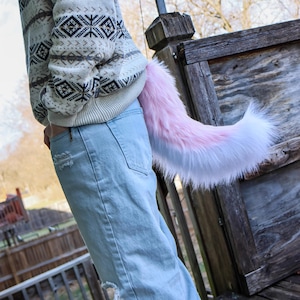 May include: A fluffy, pink and white faux fur tail, attached to the back of light blue jeans. The tail is curved and has a gradient of colors, transitioning from pink to white. The person is wearing a cream-colored sweater with a black and white geometric pattern.