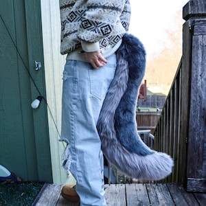 May include: A faux fur tail in shades of gray and blue, attached to the back of light blue jeans. The tail is long and fluffy, with a gradient effect from dark to light gray. The person is wearing a patterned sweater.