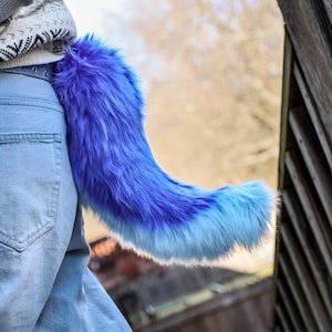 May include: A blue and light blue faux fur tail attached to a belt loop. The tail is a gradient of dark to light blue, with a fluffy texture. The tail is attached to a person wearing blue jeans.