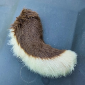 Puede incluir: Un accesorio de cola de piel sintética curvada y afelpada con un diseño de dos tonos. La sección superior es de un color marrón intenso y profundo, mientras que la parte inferior y la punta redondeada son de un blanco crema contrastante.