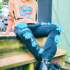 May include: Dark wash denim jeans with distressed details and turquoise faux fur accents. The jeans are paired with a tan sweatshirt featuring the word "Cruisin" and a colorful graphic. The outfit is completed with black boots.
