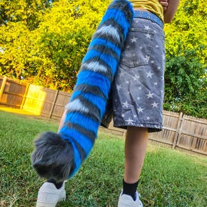 May include: A long, plush, faux fur tail featuring alternating horizontal stripes of vibrant blue, dark gray, and white. The tail has a thick, rounded tip and is attached to a blue belt worn over gray shorts with a white star pattern and white sneakers.