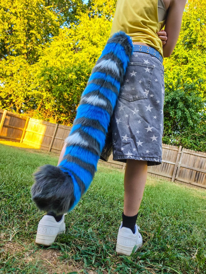 May include: A long, plush, faux fur tail featuring alternating horizontal stripes of vibrant blue, dark gray, and white. The tail has a thick, rounded tip and is attached to a blue belt worn over gray shorts with a white star pattern and white sneakers.