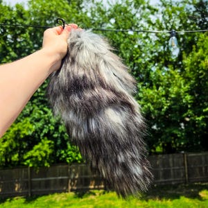 Könnte beinhalten: Ein langer, flauschiger Kunstfellschwanz mit einem variierenden Farbmuster aus Anthrazit, Schwarz und Weiß. Der Schwanz hat eine spitz zulaufende Form und verfügt über einen kleinen schwarzen Metallclip zur Befestigung mit dichter Textur.