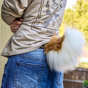 May include: A fluffy, faux fur tail in white and brown, attached to the back of blue jeans. The tail has a brown patch at the base and transitions to white. The person is wearing a beige sweatshirt with the word "MIRACLE" printed on the back.